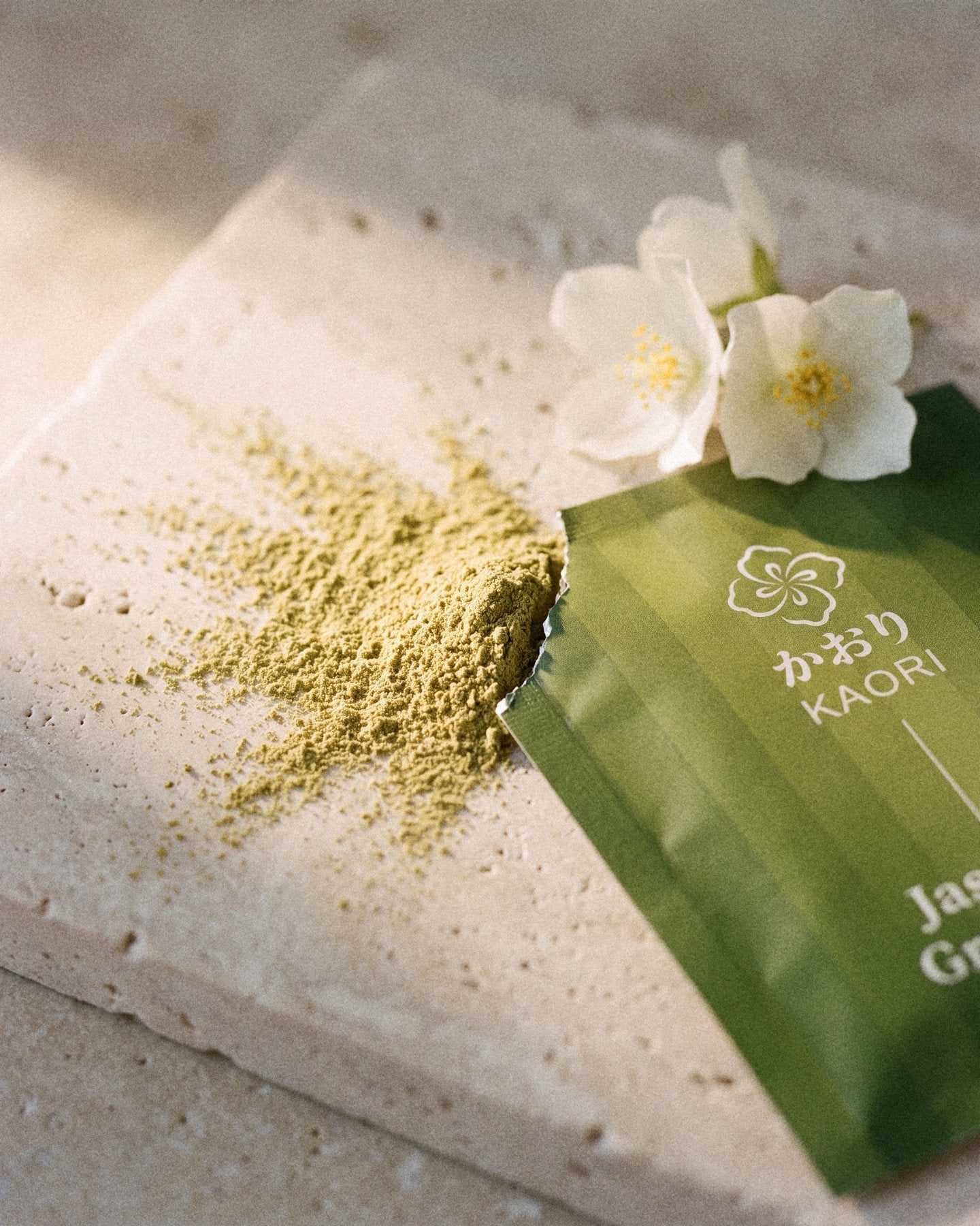 A torn Jasmine Green Tea sachet on pale travertine, its powder spilling out beside two jasmine blossoms