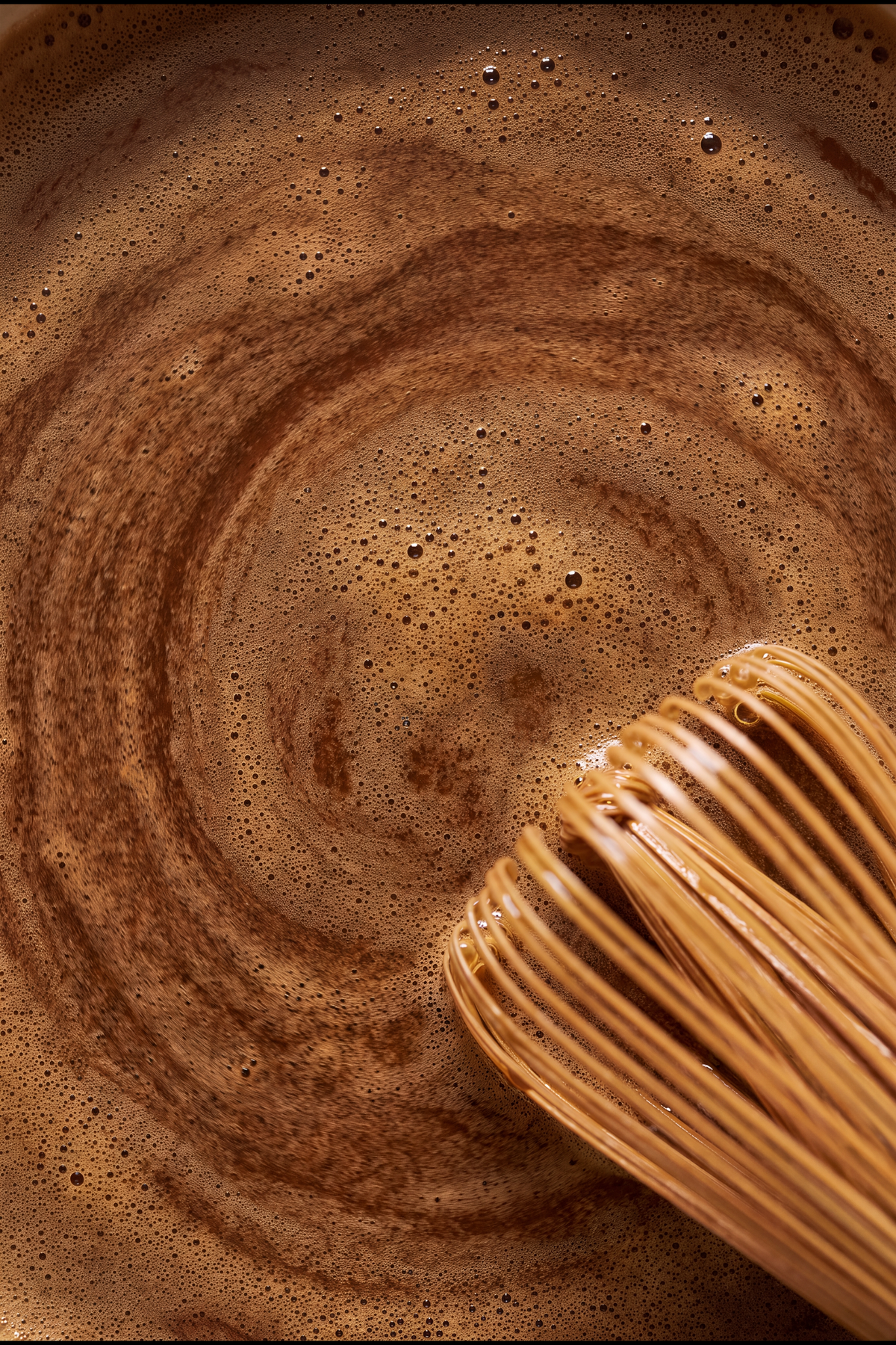 A bamboo whisk drawing circles through the foam of a hojicha latte