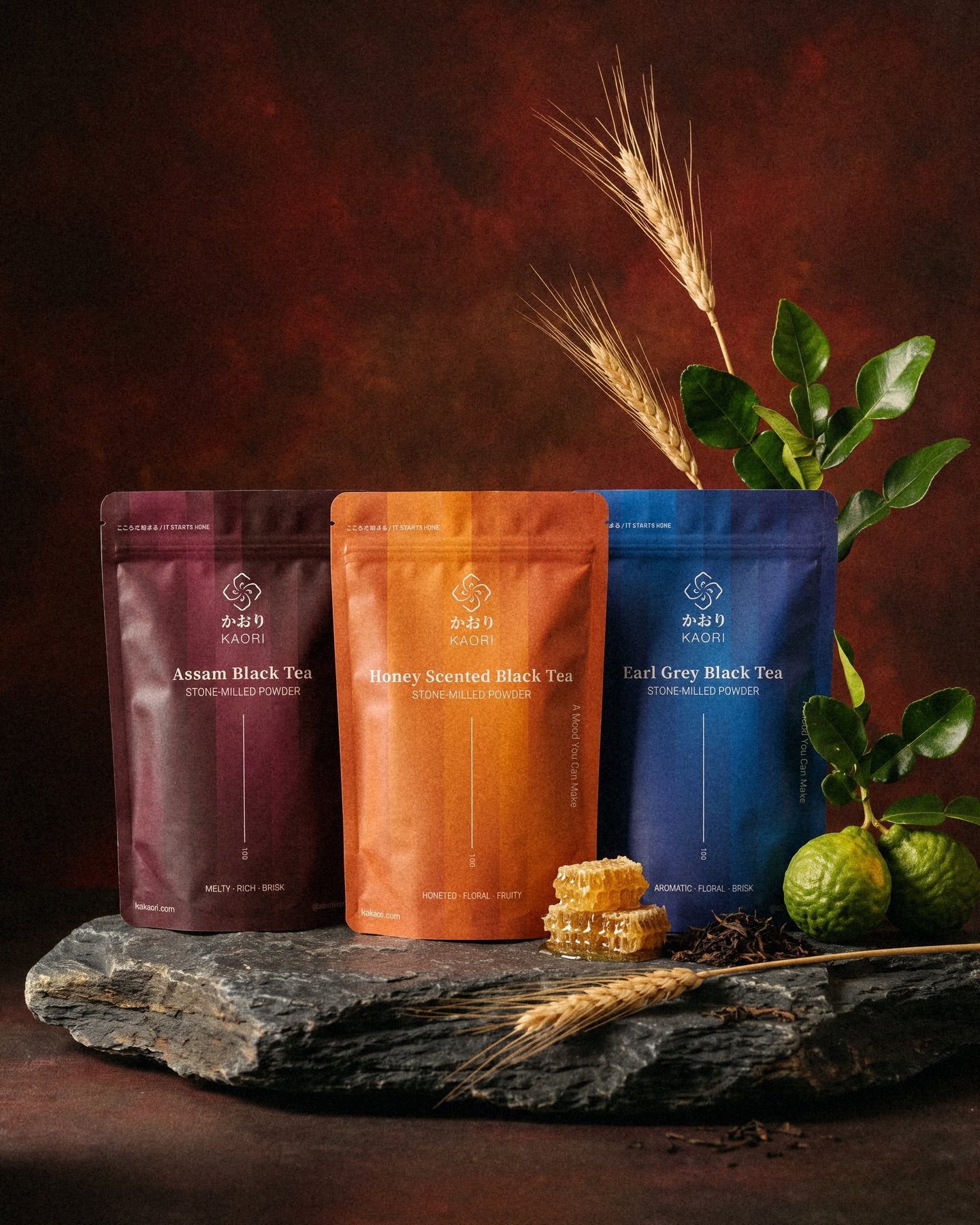 Assam, Honey Scented and Earl Grey pouches standing on dark slate with honeycomb, bergamot and wheat, before an oxblood painted backdrop