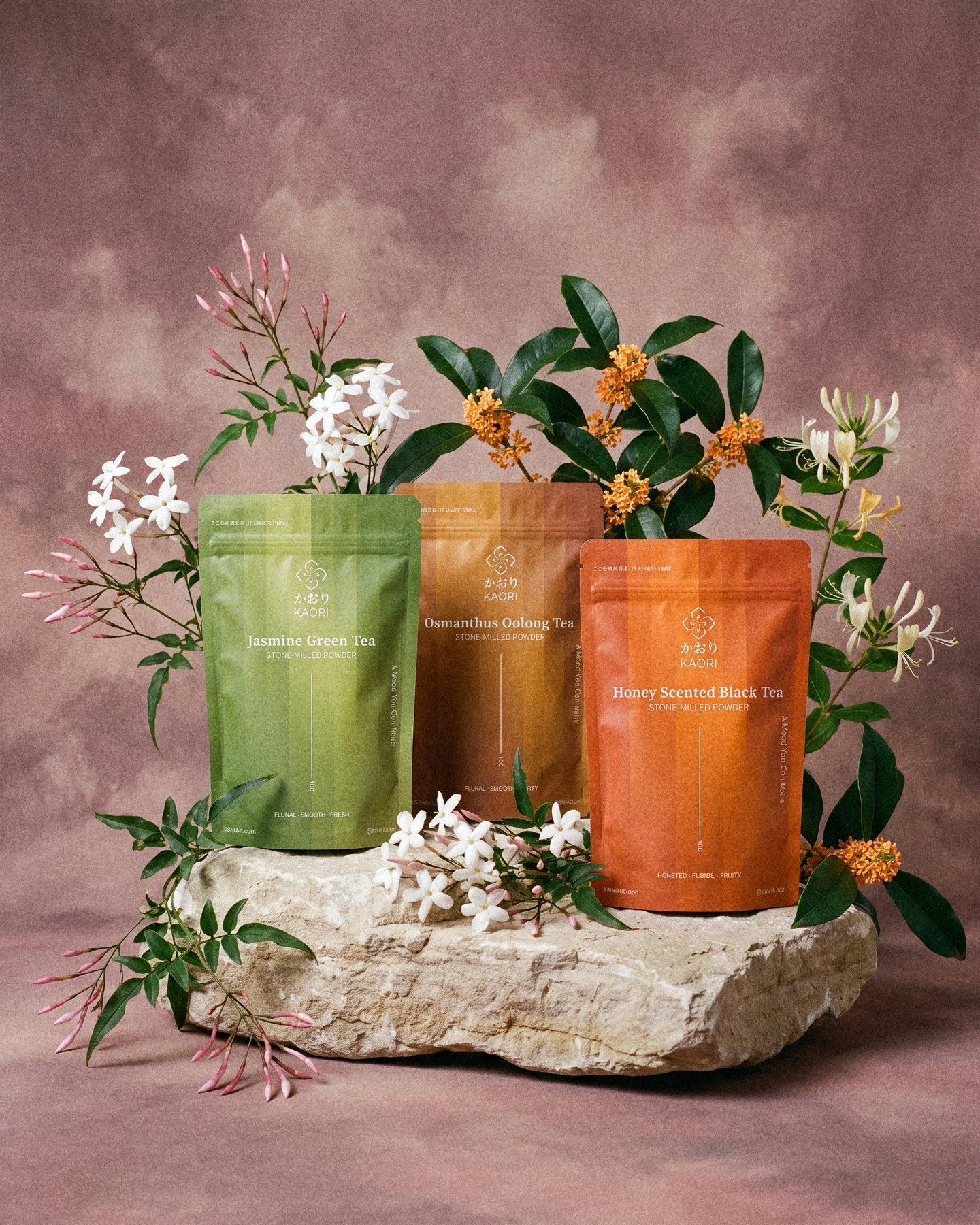 Jasmine Green Tea, Osmanthus Oolong and Honey Scented Black Tea pouches standing on a pale limestone boulder among jasmine, osmanthus and honeysuckle, before a dusty rose painted backdrop