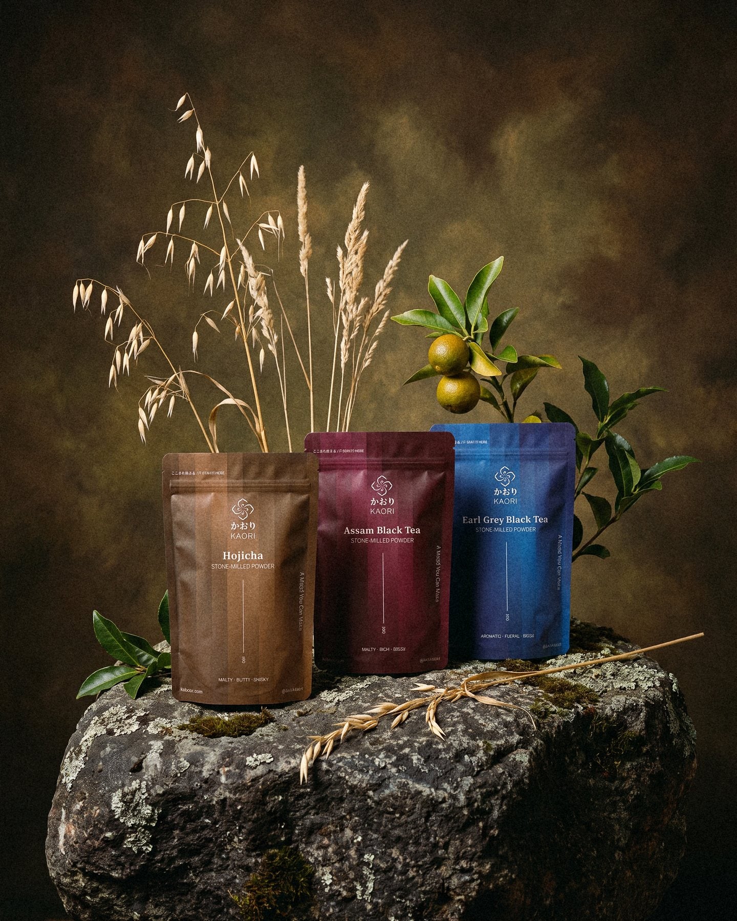 Hojicha, Assam and Earl Grey pouches standing on a lichen-flecked granite rock among wild oats and a bergamot branch, before a deep olive painted backdrop