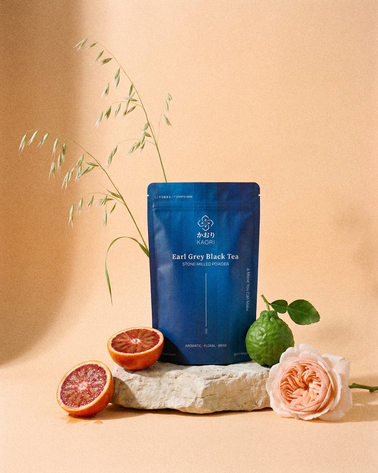 An Earl Grey Black Tea pouch on a pale stone against apricot paper, halved blood oranges, a bergamot and a garden rose at its foot