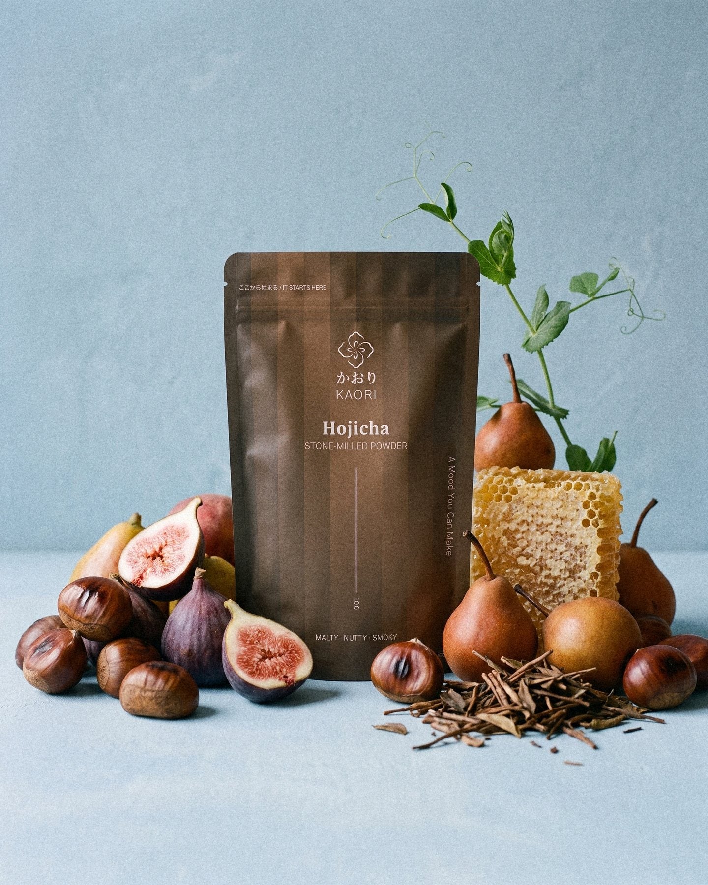 A Hojicha pouch among split figs, chestnuts, pears and honeycomb against powder-blue plaster